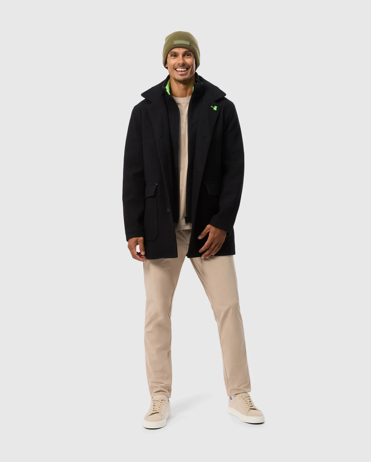 A person is smiling, wearing the Psycho Bunny MENS STAFFORD WOOL BLEND COAT in black over tan pants and beige sneakers, topped with an olive green beanie against a plain gray background.