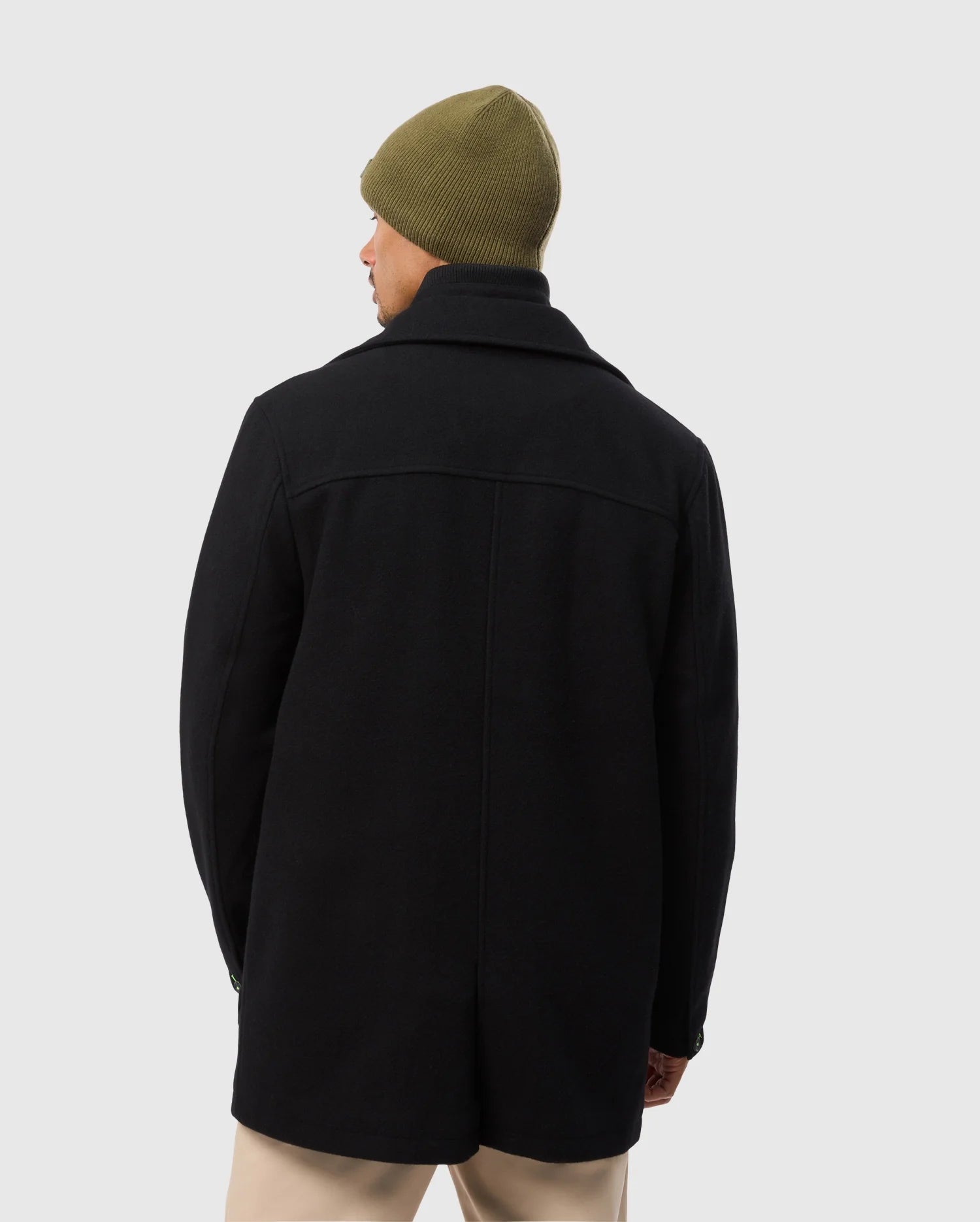A person wearing a Psycho Bunny MENS STAFFORD WOOL BLEND COAT in black and an olive green beanie stands with their back to the camera, paired with beige pants against a plain light gray background.
