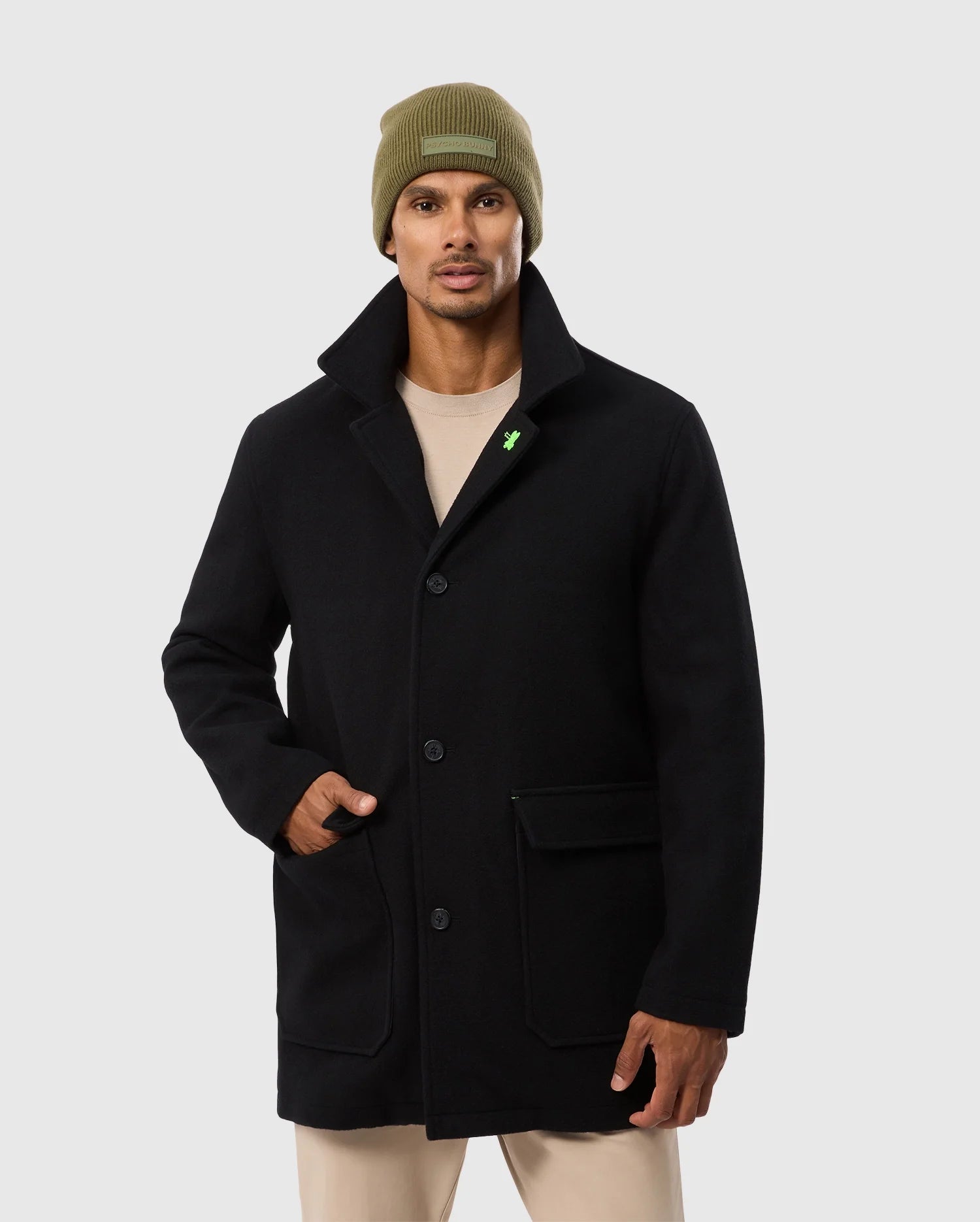 A man with a mustache in the Psycho Bunny MENS STAFFORD WOOL BLEND COAT, B6N551D200, paired with an olive beanie. One hand is in his pocket; the jacket has large buttons and a small green pin against a plain gray background.