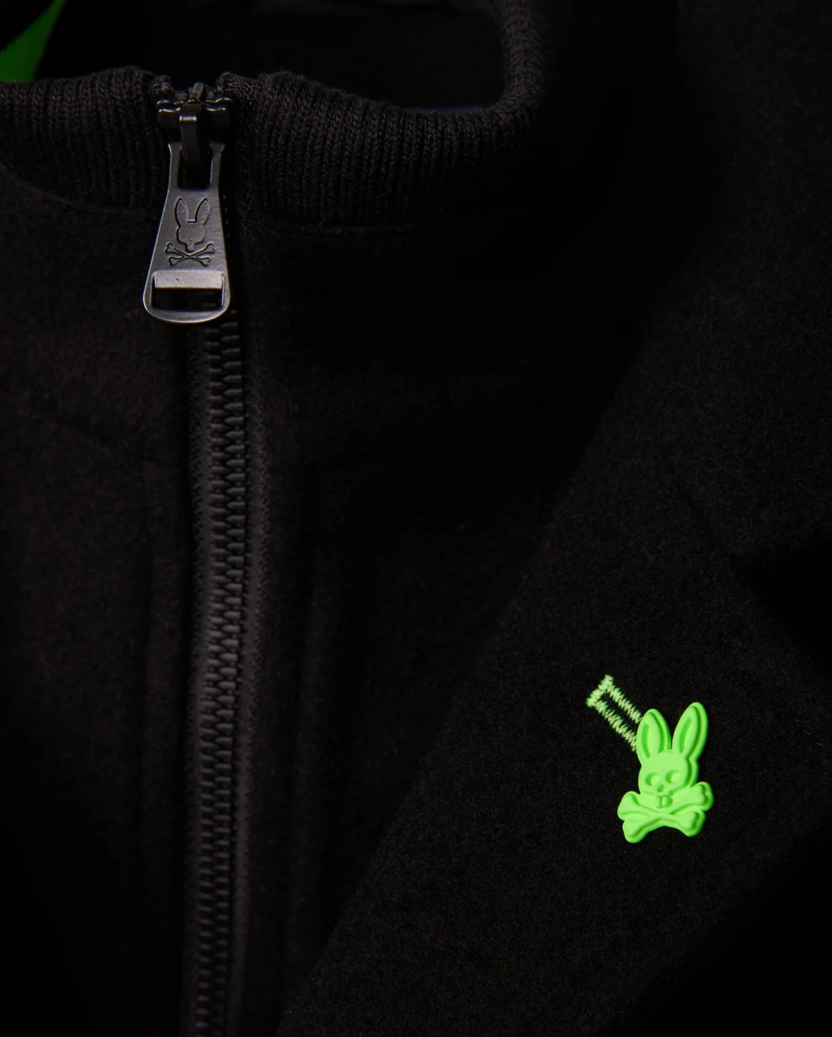 A close-up of the Psycho Bunny MENS STAFFORD WOOL BLEND COAT - B6N551D200 shows a partially open zipper revealing a green bunny skull and crossbones emblem on the right side. The zipper pull also features the brand's logo, highlighting the textures of this stylish black men's winter coat.