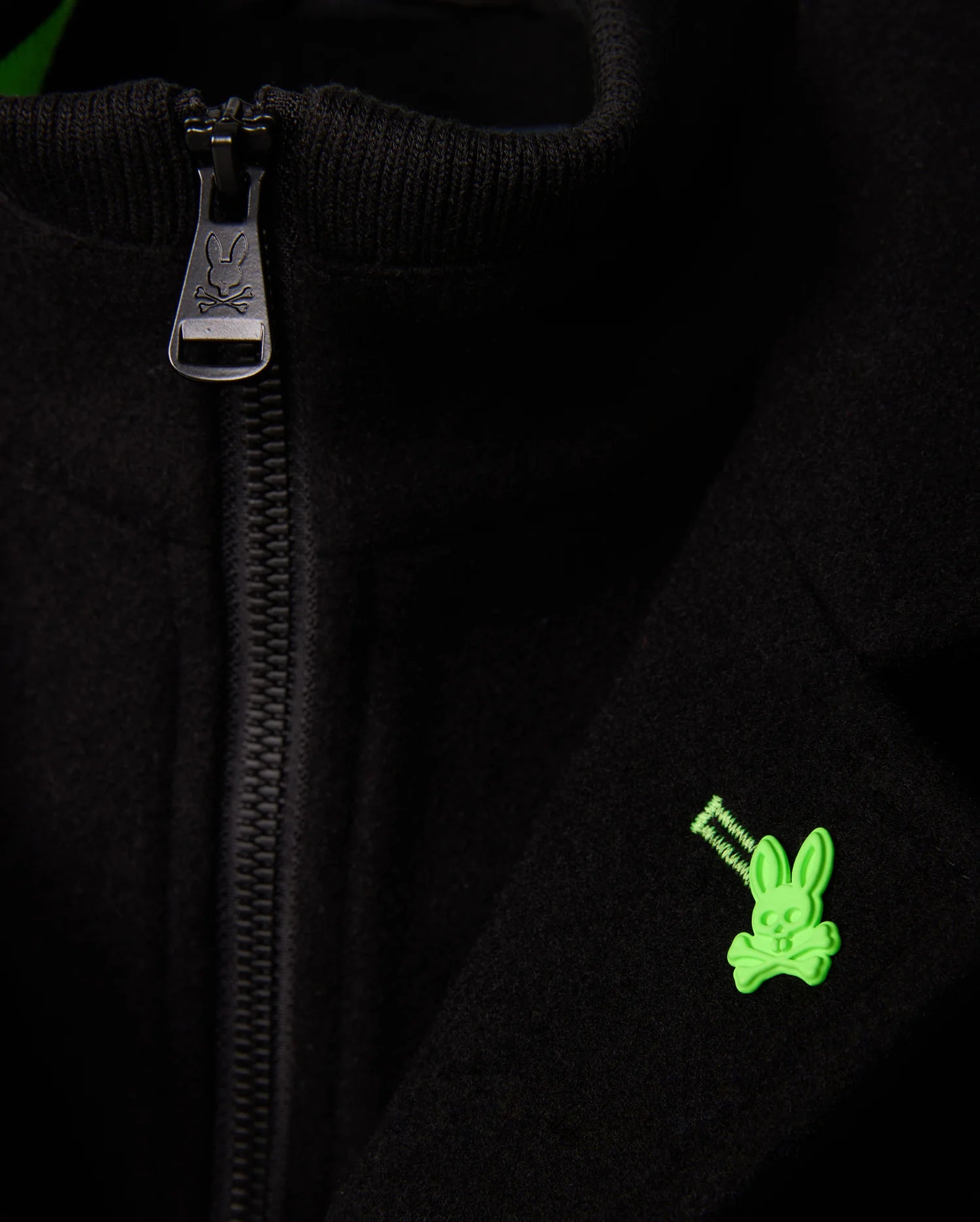 A close-up of the Psycho Bunny MENS STAFFORD WOOL BLEND COAT - B6N551D200 shows a partially open zipper revealing a green bunny skull and crossbones emblem on the right side. The zipper pull also features the brand's logo, highlighting the textures of this stylish black men's winter coat.