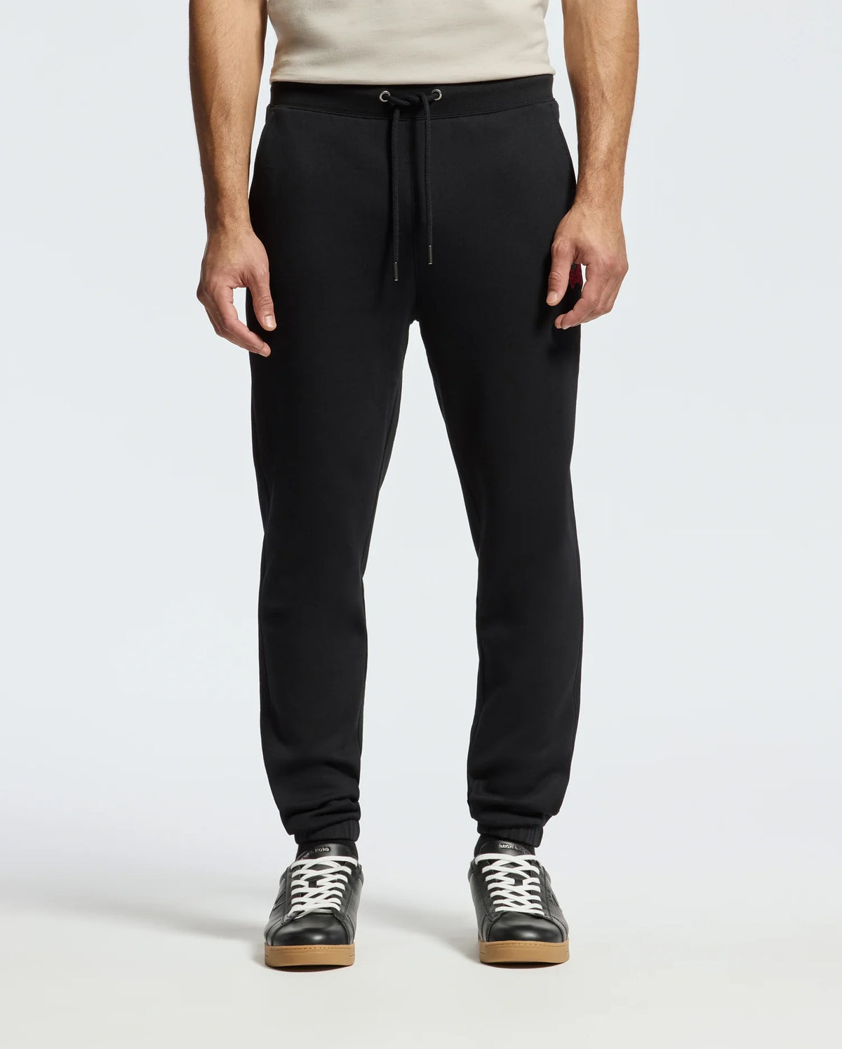 A person is wearing the Psycho Bunny MENS PIERCE ESSENTIAL FRENCH TERRY SWEATPANT in sleek black, complete with a drawstring waist, paired with black sneakers featuring white laces and soles. The background is plain white, and the upper body is partially visible.