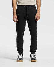 A person is wearing the Psycho Bunny MENS PIERCE ESSENTIAL FRENCH TERRY SWEATPANT in sleek black, complete with a drawstring waist, paired with black sneakers featuring white laces and soles. The background is plain white, and the upper body is partially visible.
