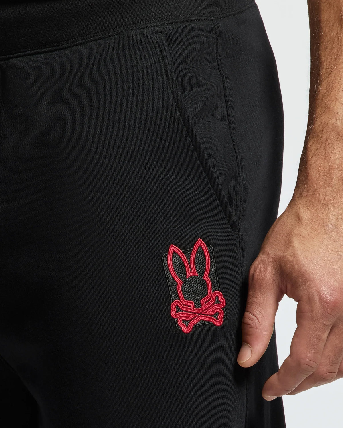 Close-up of a person's hand resting near the pocket of their black MENS PIERCE ESSENTIAL FRENCH TERRY SWEATPANT - B6P136E200 by Psycho Bunny, featuring a red embroidered logo of a bunny with crossbones.