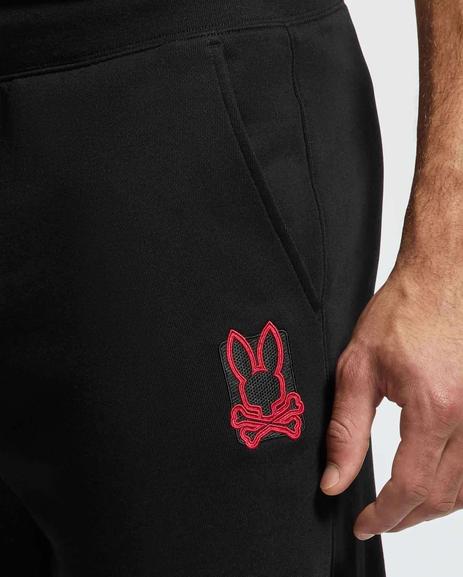 Close-up of a person's hand resting near the pocket of their black MENS PIERCE ESSENTIAL FRENCH TERRY SWEATPANT - B6P136E200 by Psycho Bunny, featuring a red embroidered logo of a bunny with crossbones.