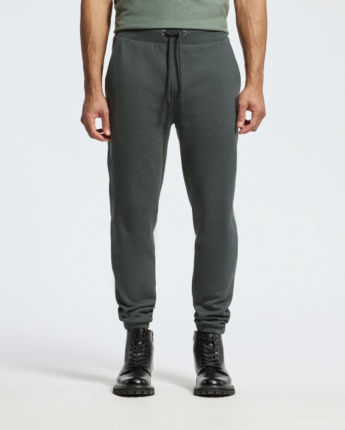 A person is wearing the MENS PIERCE ESSENTIAL FRENCH TERRY SWEATPANT (B6P136E200) by Psycho Bunny, which are dark green joggers made from Cotton French Terry and feature a drawstring waist. They are paired with black lace-up boots. The upper body is partially visible, displaying a green shirt against a plain light gray background.