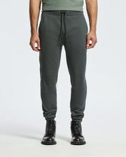 A person is wearing the MENS PIERCE ESSENTIAL FRENCH TERRY SWEATPANT (B6P136E200) by Psycho Bunny, which are dark green joggers made from Cotton French Terry and feature a drawstring waist. They are paired with black lace-up boots. The upper body is partially visible, displaying a green shirt against a plain light gray background.