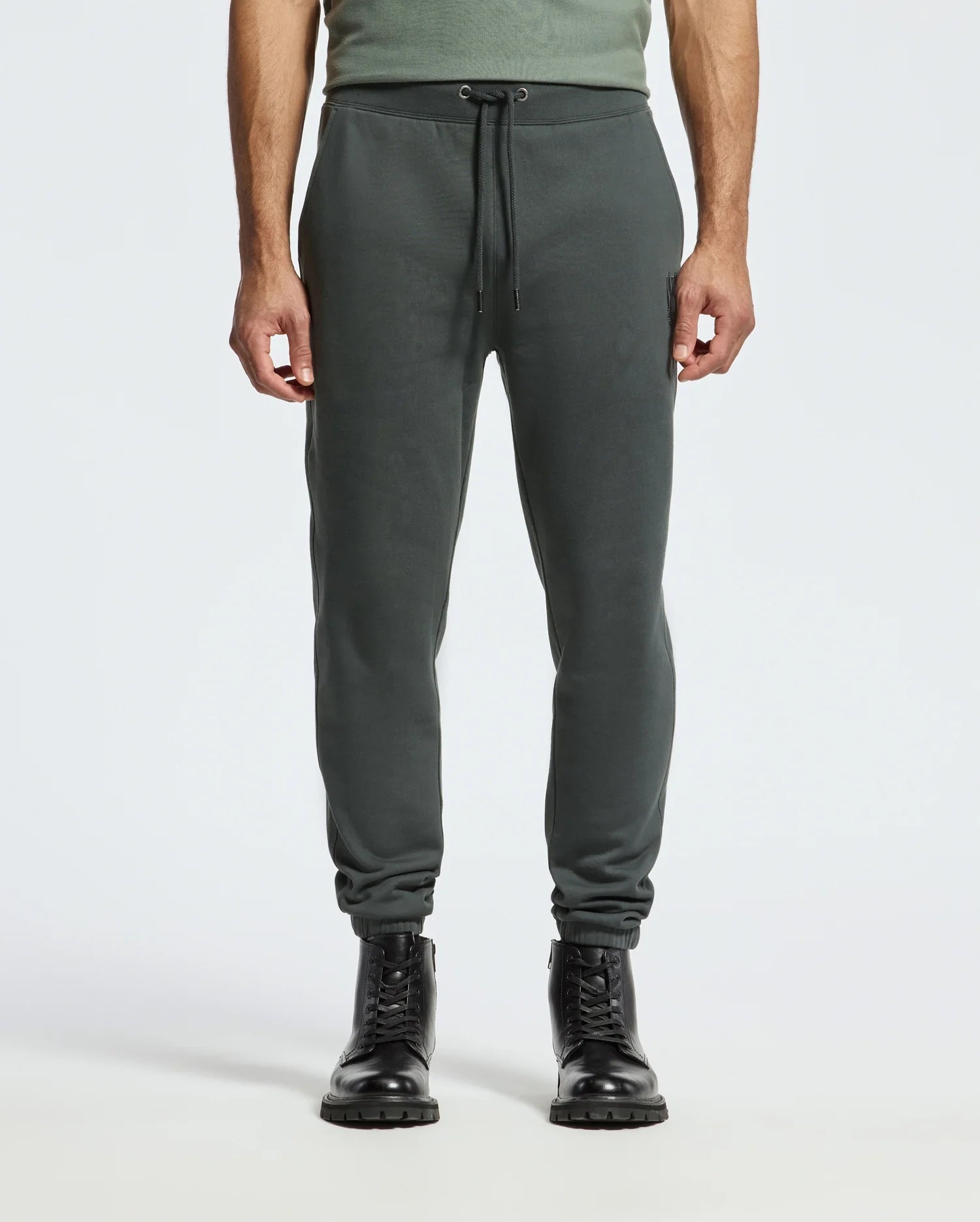A person is wearing the MENS PIERCE ESSENTIAL FRENCH TERRY SWEATPANT (B6P136E200) by Psycho Bunny, which are dark green joggers made from Cotton French Terry and feature a drawstring waist. They are paired with black lace-up boots. The upper body is partially visible, displaying a green shirt against a plain light gray background.