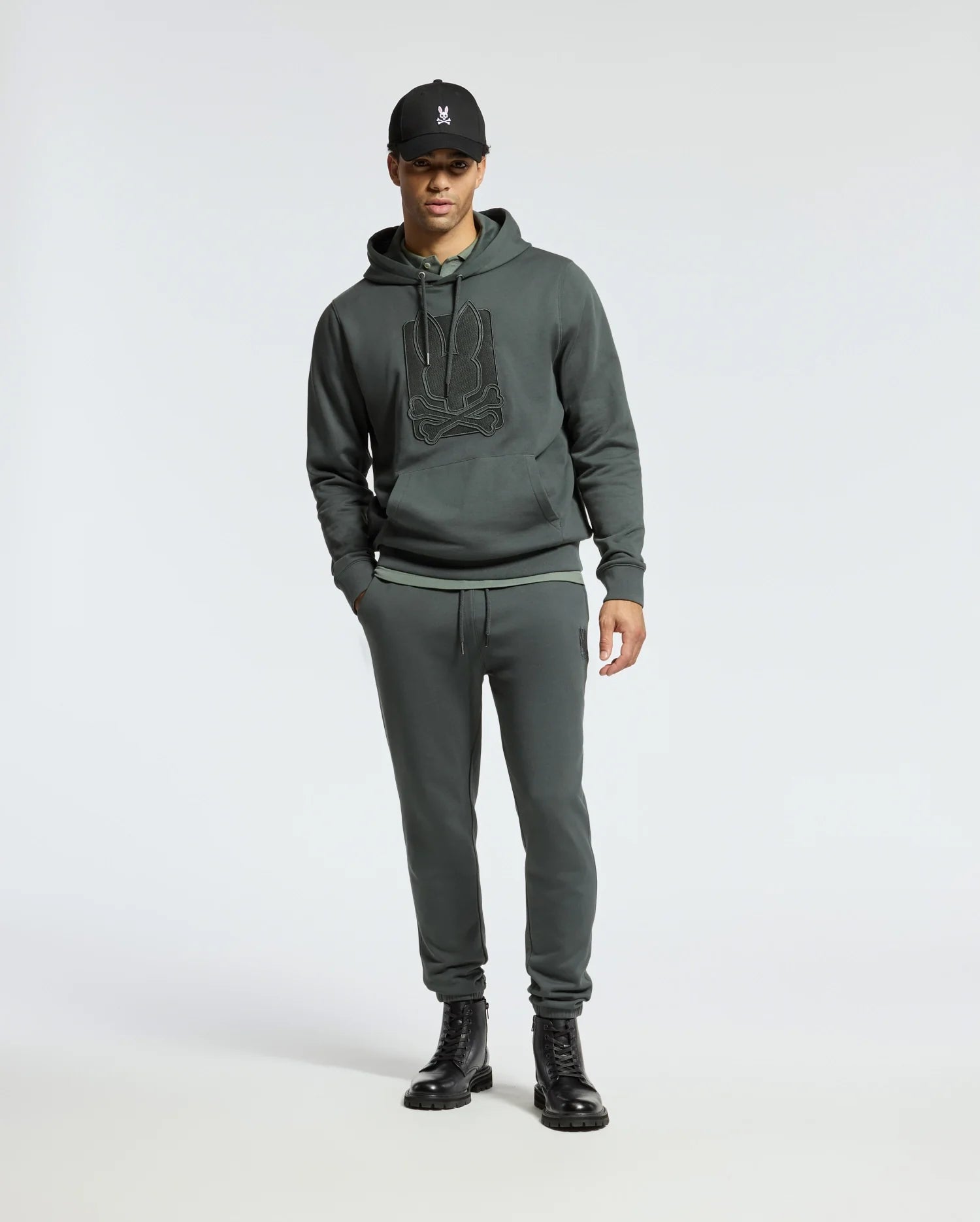 A person wearing a dark hoodie with a logo, Psycho Bunny MENS PIERCE ESSENTIAL FRENCH TERRY SWEATPANT - B6P136E200, black boots, and a black cap stands against a plain, light background.