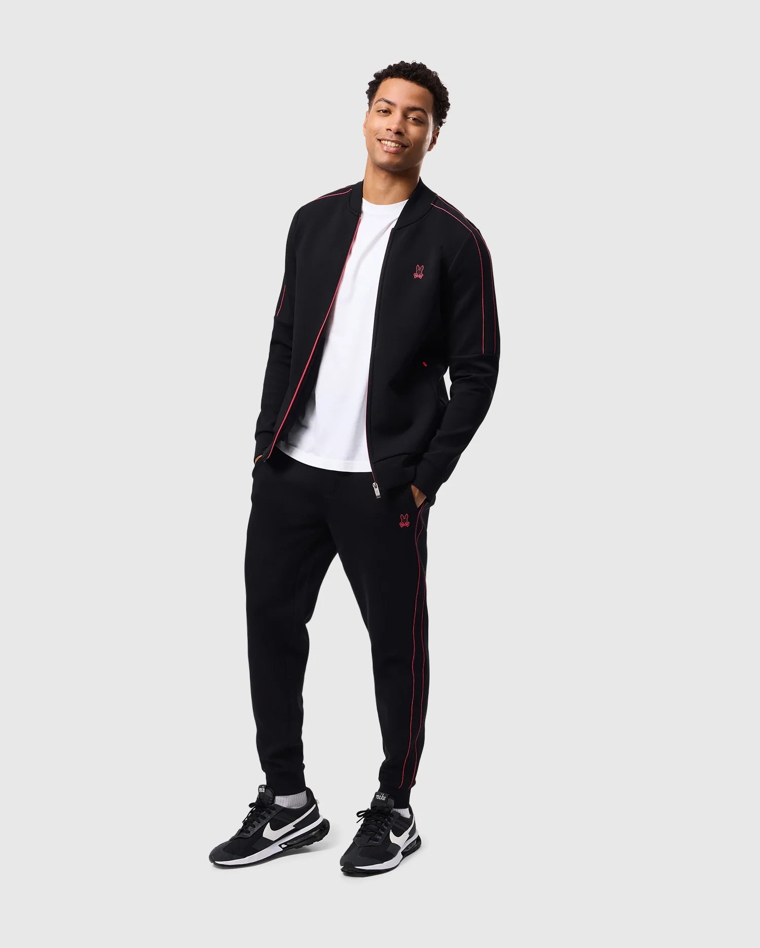 A man is standing and smiling while dressed in a black Psycho Bunny MENS TRAVIS TRACK PANT - B6P143D200 with red accents and a white T-shirt, made of high-quality cotton-blend for regular fit. He is wearing black and white sneakers. The background is plain and light-colored.