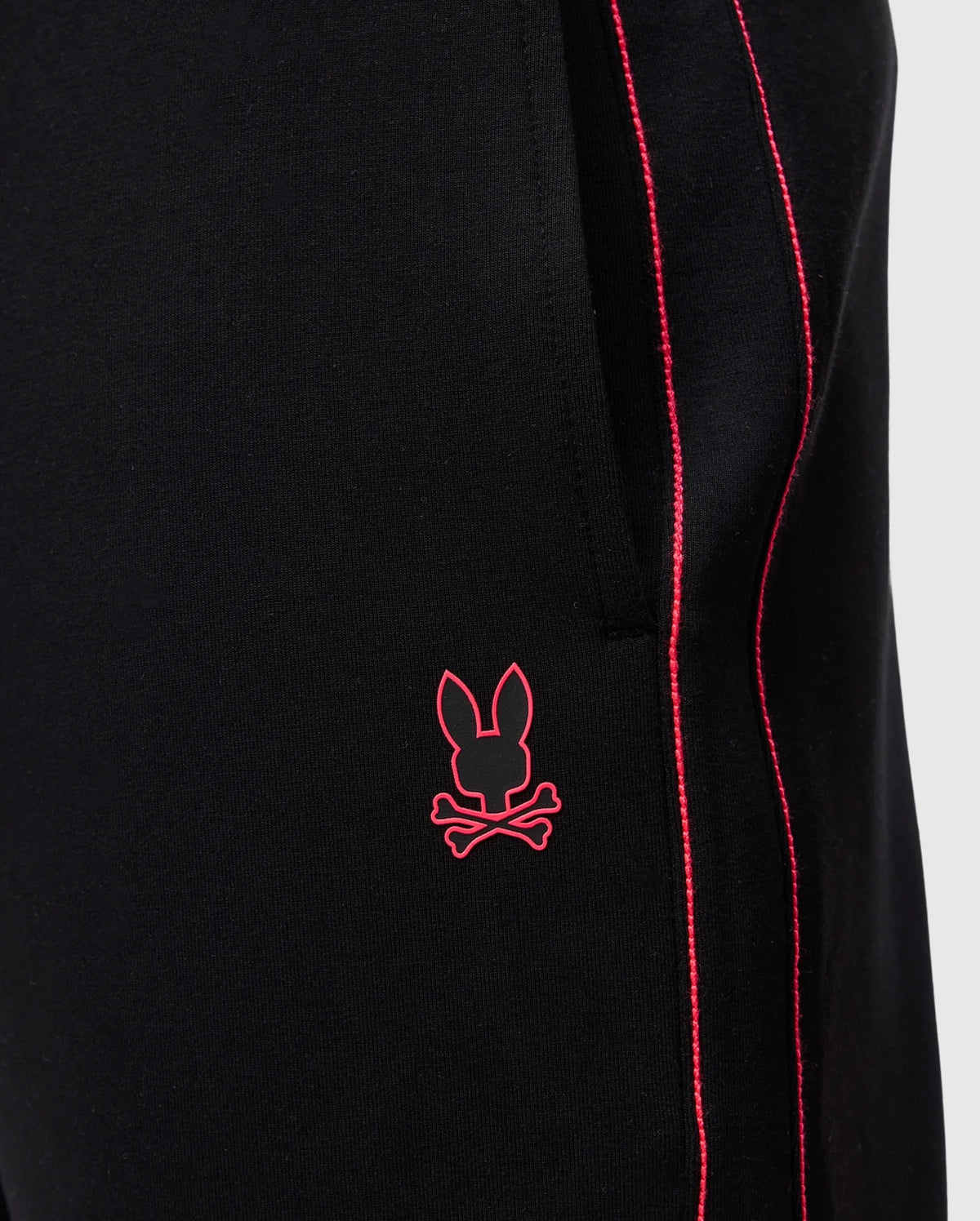 Close-up of Psycho Bunny's MENS TRAVIS TRACK PANT - B6P143D200 in black, featuring red piping along the side seam, a silicone heat transfer bunny near the pocket, and crafted from high-quality cotton-blend fabric.
