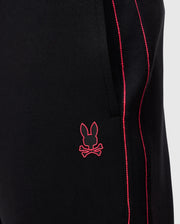 Close-up of Psycho Bunny's MENS TRAVIS TRACK PANT - B6P143D200 in black, featuring red piping along the side seam, a silicone heat transfer bunny near the pocket, and crafted from high-quality cotton-blend fabric.