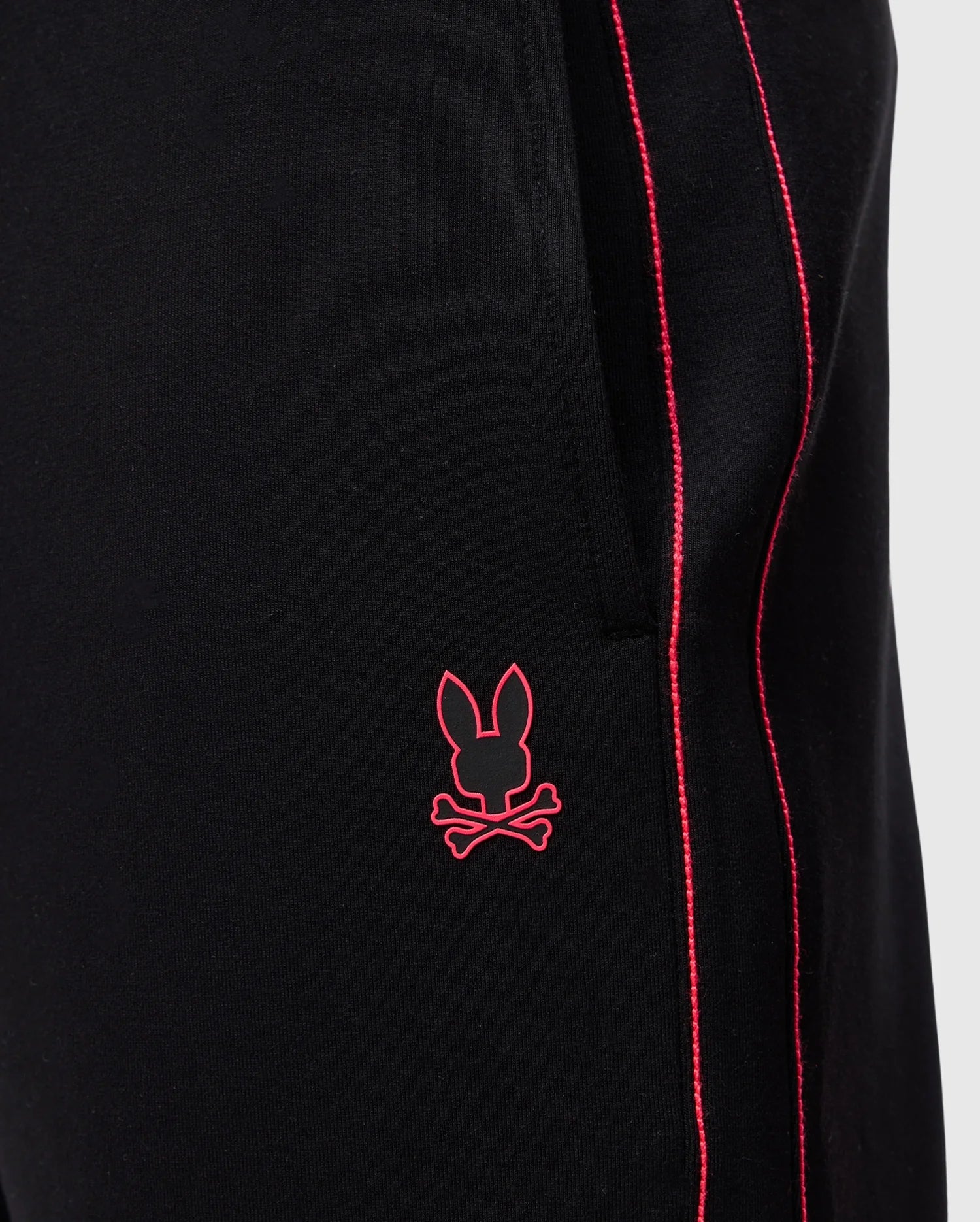 Close-up of Psycho Bunny's MENS TRAVIS TRACK PANT - B6P143D200 in black, featuring red piping along the side seam, a silicone heat transfer bunny near the pocket, and crafted from high-quality cotton-blend fabric.