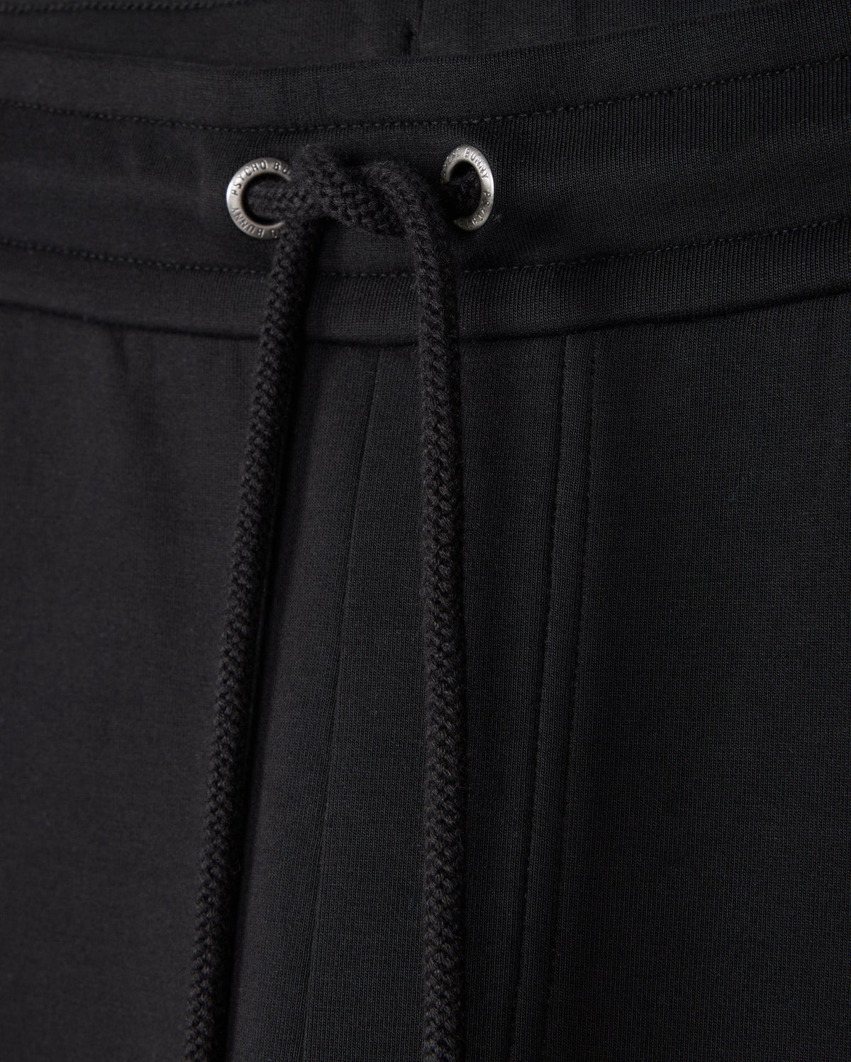 Close-up of black Psycho Bunny MENS TRAVIS TRACK PANT - B6P143D200 featuring a drawstring waistband. The drawstrings are threaded through metal eyelets, and the high-quality cotton-blend fabric has a smooth, slightly textured appearance. The stitching and seams are visible, showcasing the garment's craftsmanship.