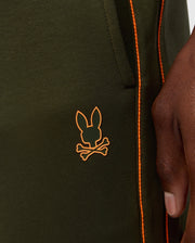 Close-up of a person wearing MENS TRAVIS TRACK PANT - B6P143D200 by Psycho Bunny, in green with orange piping along the seams. The high-quality cotton-blend pants feature invisible zip pockets and a logo of a stylized bunny head and crossbones in orange, located near the pocket. The person's hand rests beside the pocket.