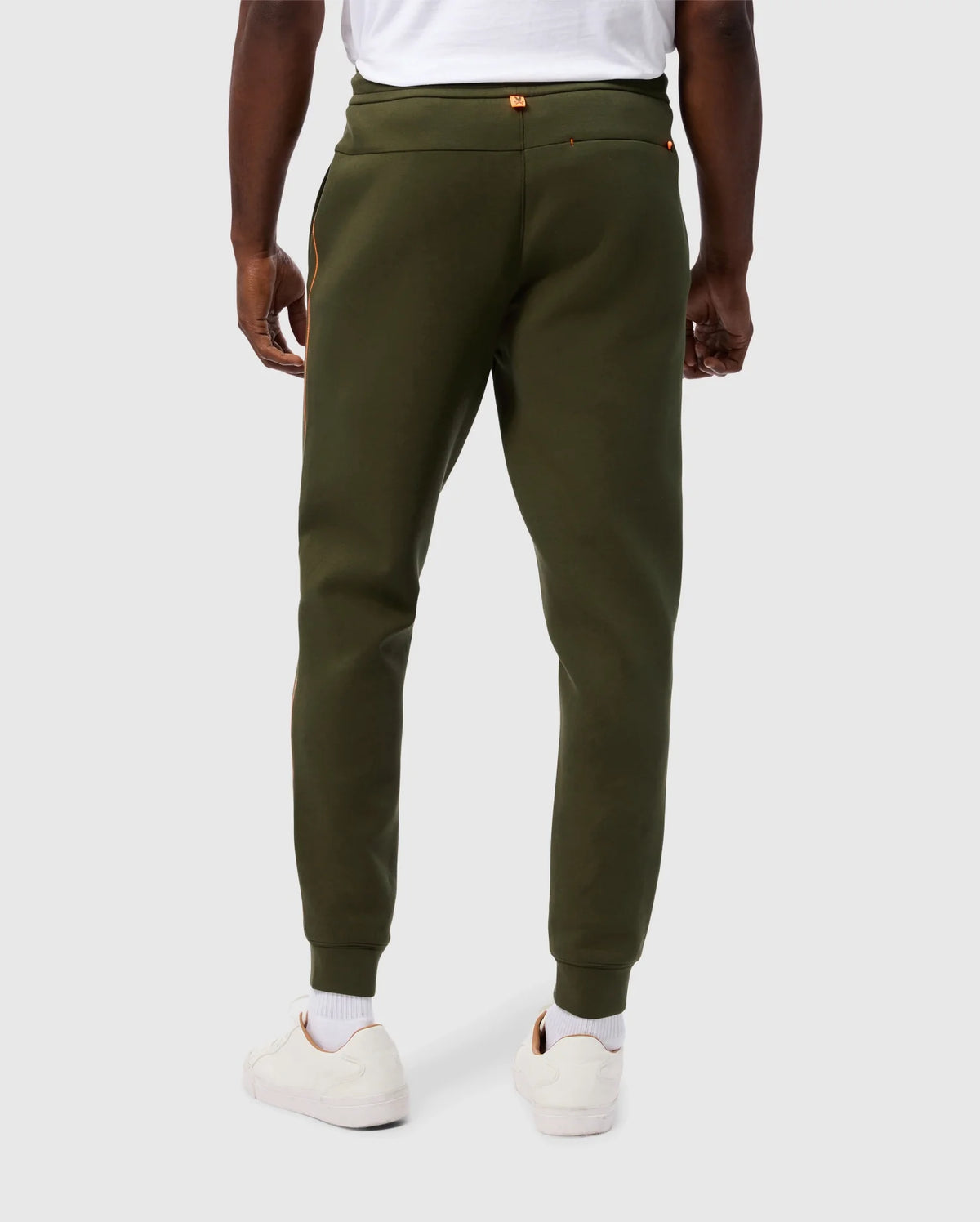 Rear view of a person wearing olive green Psycho Bunny MENS TRAVIS TRACK PANT - B6P143D200 and a white T-shirt. The high-quality cotton-blend joggers feature invisible zip pockets with gold zippers. The person is dressed in white sneakers and white socks, set against a plain, light-colored background.