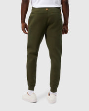 Rear view of a person wearing olive green Psycho Bunny MENS TRAVIS TRACK PANT - B6P143D200 and a white T-shirt. The high-quality cotton-blend joggers feature invisible zip pockets with gold zippers. The person is dressed in white sneakers and white socks, set against a plain, light-colored background.