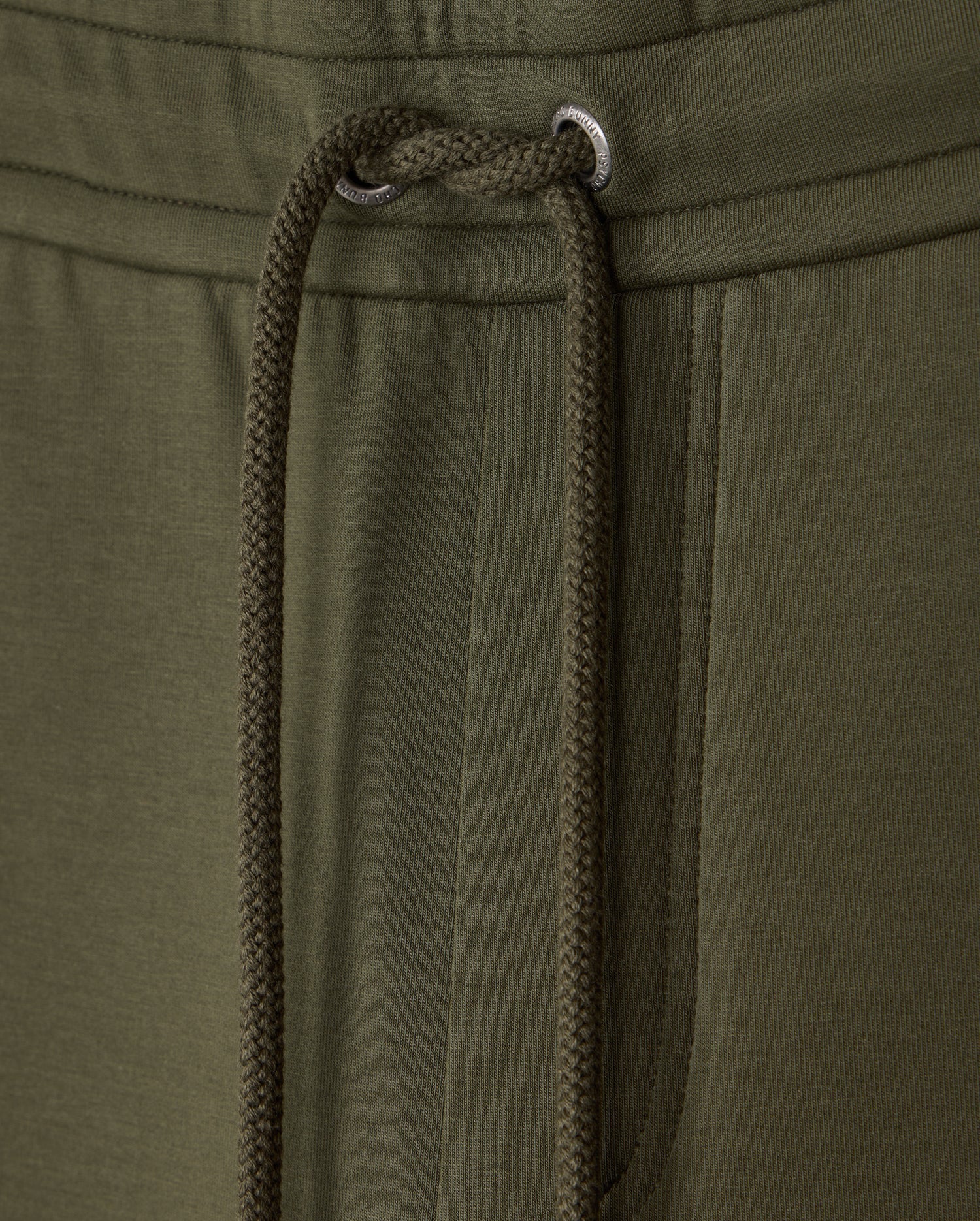 Close-up of the waistband and drawstring of a pair of olive green MENS TRAVIS TRACK PANT - B6P143D200 by Psycho Bunny. The image highlights the texture of the high-quality cotton-blend fabric and the metal eyelets through which the drawstring is threaded. A pocket is partially visible on the right side.