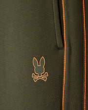 Close-up of the Psycho Bunny Men's Travis Track Pant (B6P143D200) in dark green, crafted from a high-quality cotton-blend material featuring a pocket with orange stitching. Below the pocket, there's an orange logo of a rabbit head overlaid on two crossed bones.