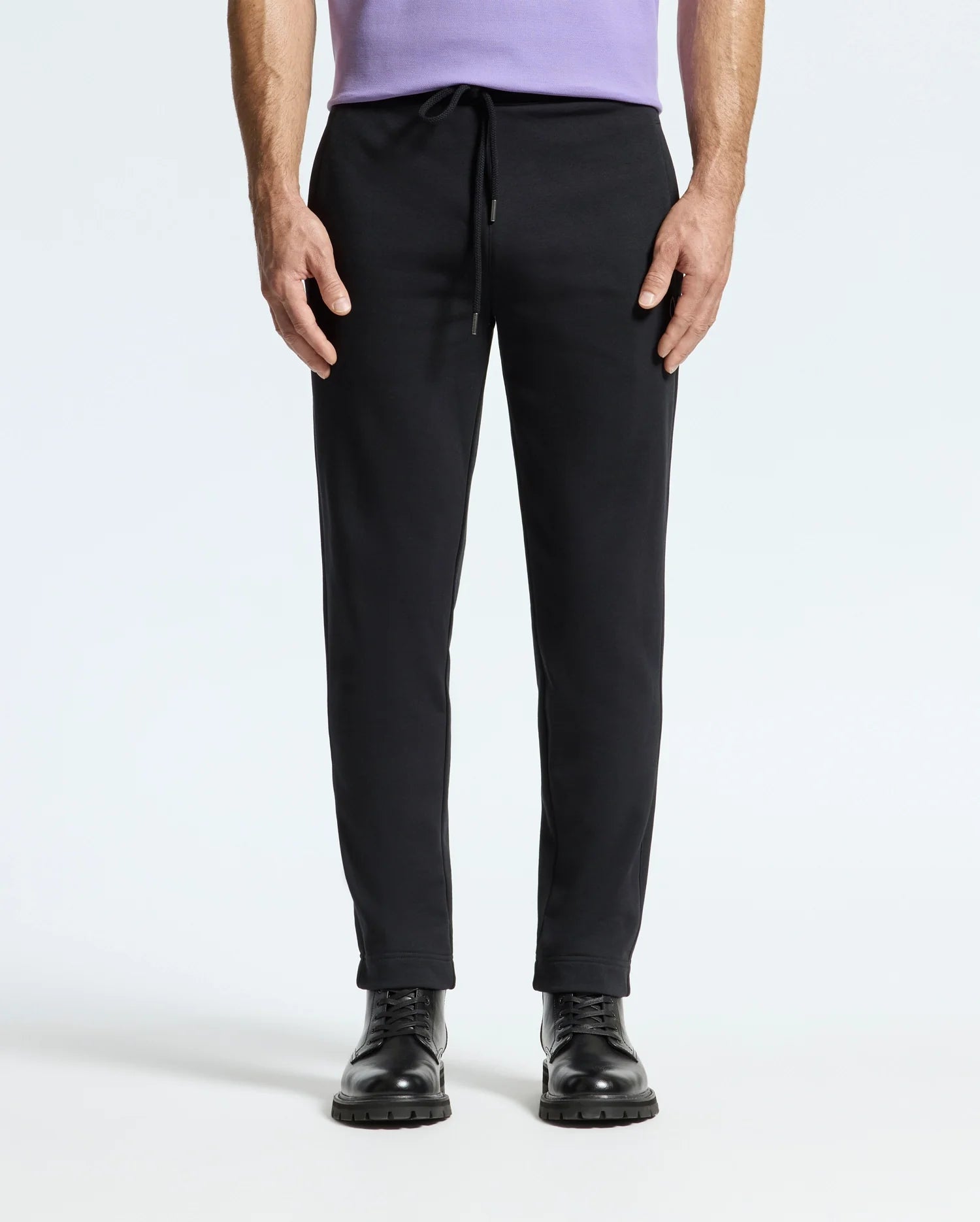 A person stands against a plain background, showcasing their outfit which includes a light purple shirt and the Psycho Bunny MENS ALEXANDER SWEATPANT - B6P155E200, complete with drawstrings. Additionally, they are wearing black lace-up boots. The focus of the image is on the lower half of their body.