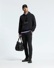 A man stands against a simple backdrop, dressed in a black hoodie featuring a graphic design, the MENS ALEXANDER SWEATPANT by Psycho Bunny, and a black cap. He is holding a large black bag and wearing black shoes.