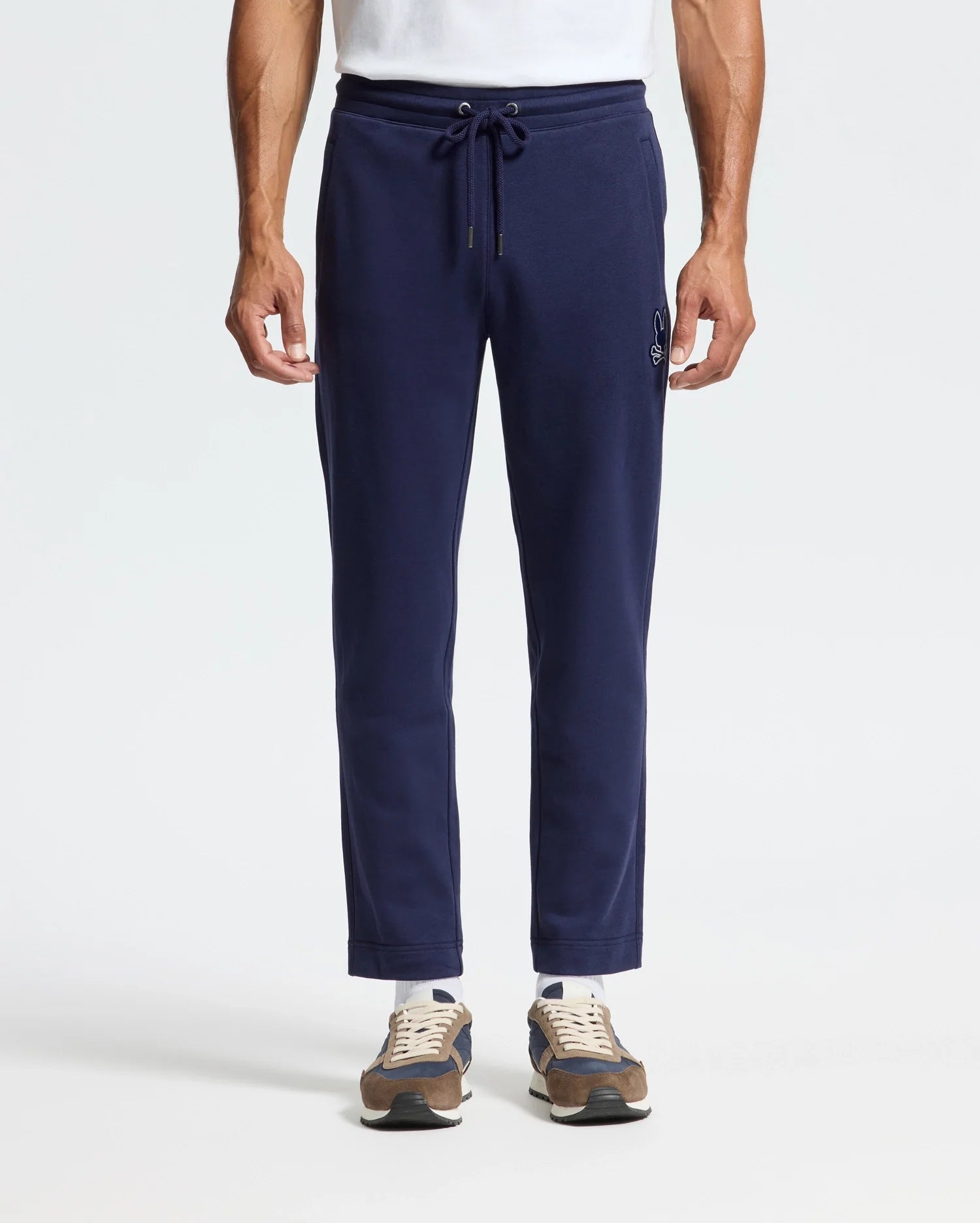 A man in Psycho Bunny's MENS ALEXANDER SWEATPANT - B6P155E200, relaxed fit navy joggers with a drawstring waist and front embroidery, paired with a white T-shirt and brown sneakers.