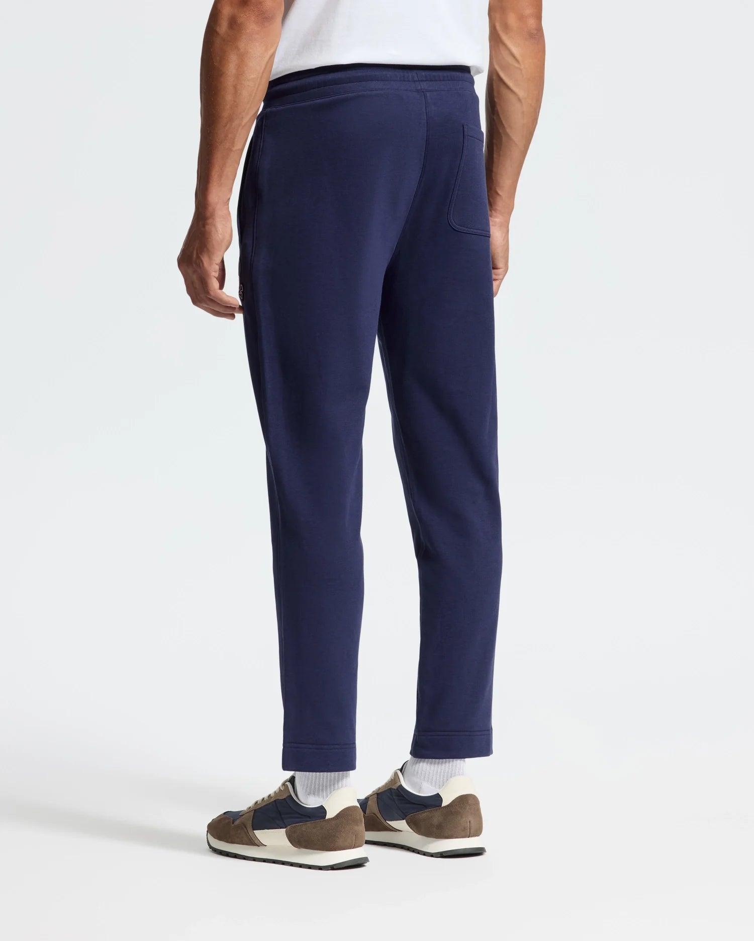 A person stands against a plain background, showcasing their lower half in Psycho Bunny's MENS ALEXANDER SWEATPANT - B6P155E200 in dark blue, with a relaxed fit and stylishly paired with brown and white sneakers.
