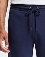 The individual sports navy blue Psycho Bunny MENS ALEXANDER SWEATPANT - B6P155E200, complete with a relaxed fit and drawstring. A white shirt is visible at the waist, and their left hand rests casually by their side.