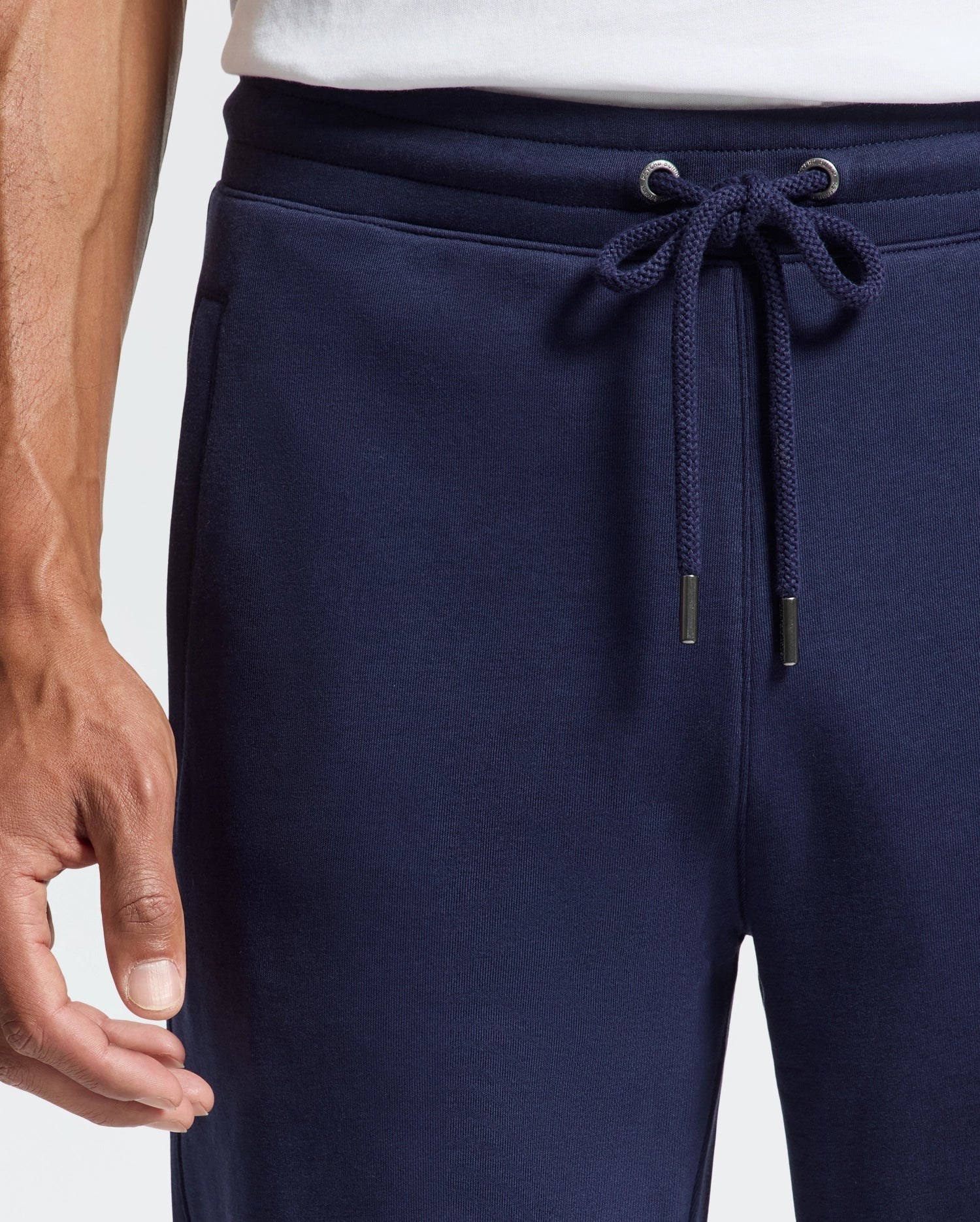 The individual sports navy blue Psycho Bunny MENS ALEXANDER SWEATPANT - B6P155E200, complete with a relaxed fit and drawstring. A white shirt is visible at the waist, and their left hand rests casually by their side.