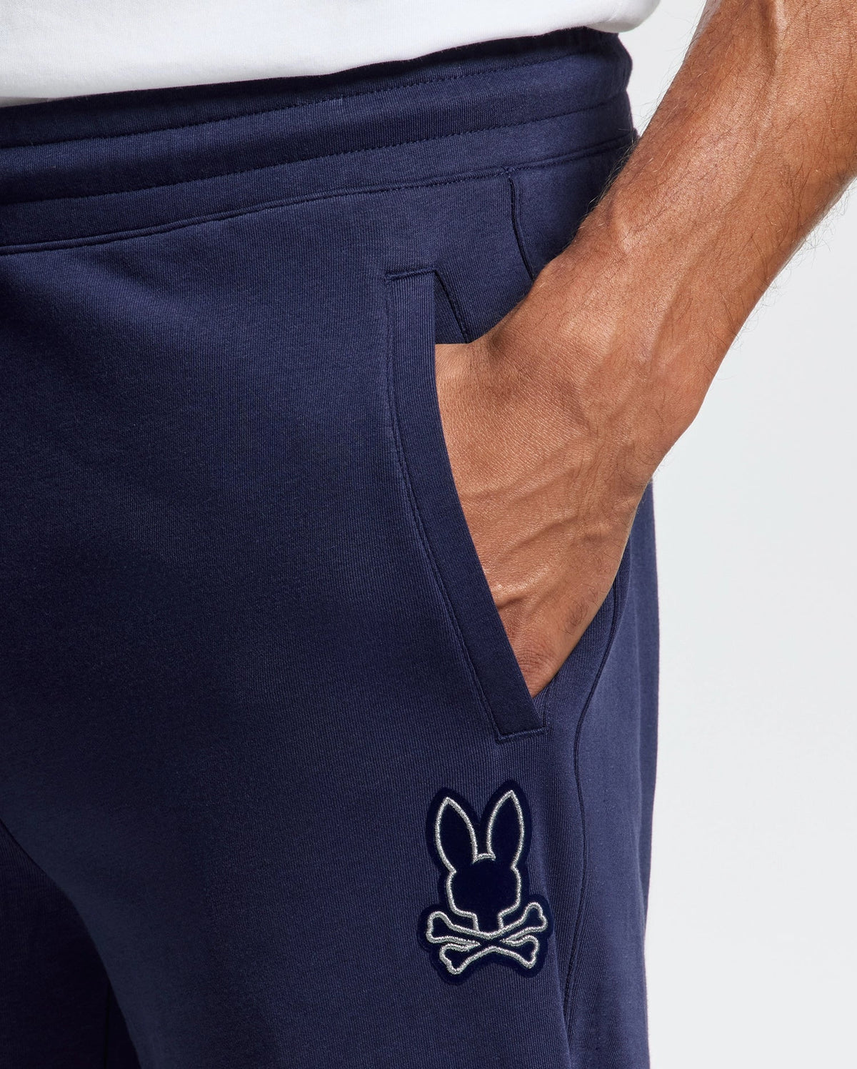 The image shows a hand in the pocket of Psycho Bunny's MENS ALEXANDER SWEATPANT - B6P155E200. These navy cotton joggers offer a relaxed fit and feature an embroidered bunny with crossbones on the left, appearing soft and comfortable.