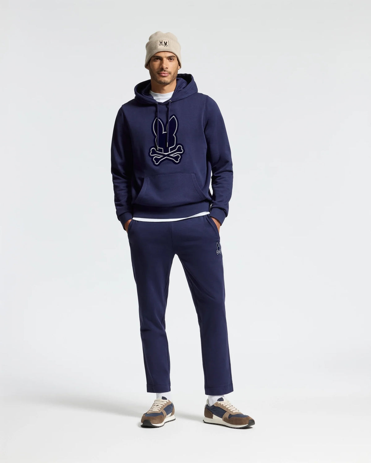 A man wearing a navy blue hoodie with a logo, Psycho Bunny's MENS ALEXANDER SWEATPANT - B6P155E200 in matching relaxed fit joggers, beige beanie, and sneakers stands against a plain background with his hands in his pockets.