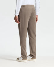 A person wearing a high-quality white long-sleeve shirt and Psycho Bunny's MENS ALEXANDER SWEATPANT - B6P155E200 in brown stands with their back to the camera. They pair this with white sneakers featuring beige soles, contributing to a relaxed look against the plain, light-colored background.