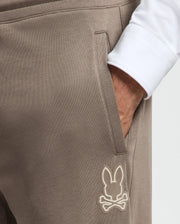 A person in MENS ALEXANDER SWEATPANT - B6P155E200 from Psycho Bunny, showcasing premium quality taupe joggers with one hand in the pocket. These cotton sweatpants feature an embroidered design of a bunny above crossed bones. The person is paired with a white long-sleeve shirt.