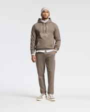 A man stands against a plain background, wearing the premium quality taupe MENS ALEXANDER SWEATPANT - B6P155E200 by Psycho Bunny, along with a hoodie featuring a cartoon face design. He layers a white shirt underneath and complements the look with casual sneakers and a beanie.