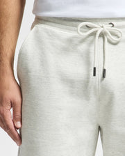A close-up features a person wearing Psycho Bunny's Men's Eldorado Brush Back Jogger (B6P285E200) in light gray with a white drawstring. Their left hand rests near their side, and they pair it with a white shirt tucked into the soft brushback cotton waistband of the joggers.