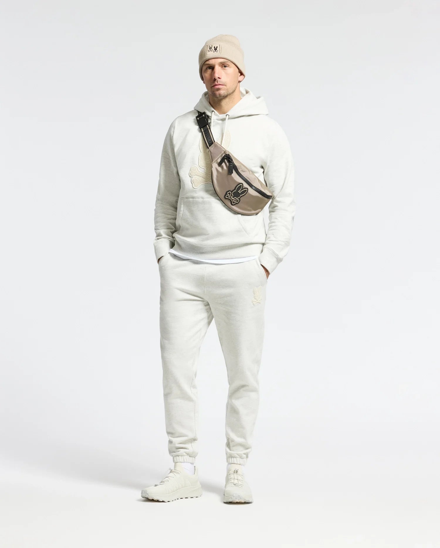 Sporting a Psycho Bunny Eldorado Brush Back Jogger, beanie, and sneakers, a person confidently stands with hands in pockets. Their crossbody bag features the Chenille Bunny logo against a plain backdrop.