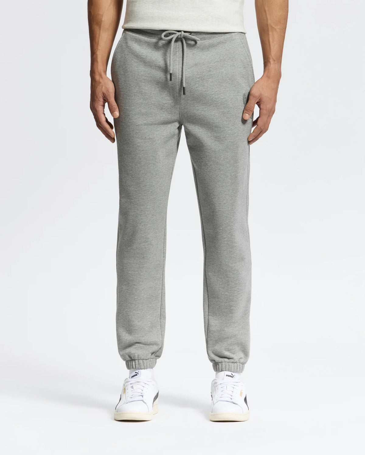 A person in sleek Psycho Bunny MENS ELDORADO BRUSH BACK JOGGER - B6P285E200, featuring a drawstring, elastic cuffs, side pockets, and brushback comfort, stands against a plain backdrop. They pair the joggers with white sneakers with beige soles.