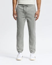 A person in sleek Psycho Bunny MENS ELDORADO BRUSH BACK JOGGER - B6P285E200, featuring a drawstring, elastic cuffs, side pockets, and brushback comfort, stands against a plain backdrop. They pair the joggers with white sneakers with beige soles.