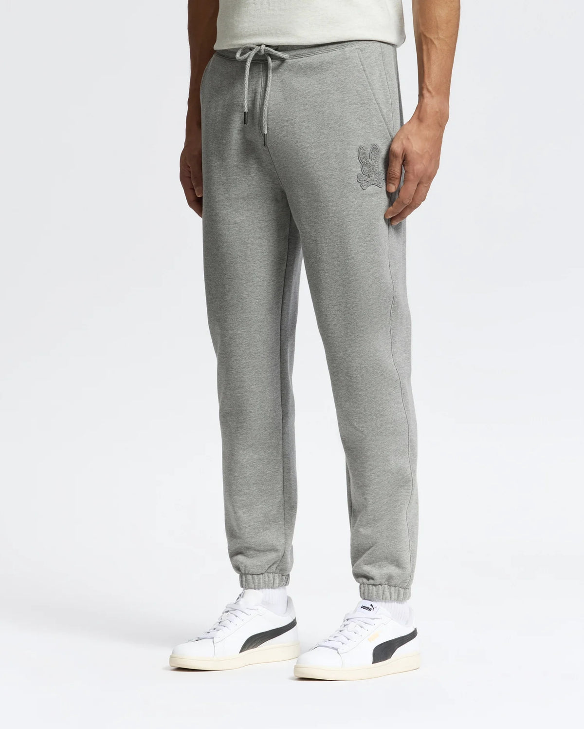 A person stands against a plain white background wearing gray Psycho Bunny's sleek MENS ELDORADO BRUSH BACK JOGGER - B6P285E200 with an embroidered design near the pocket, a white T-shirt, and white sneakers featuring black detailing.