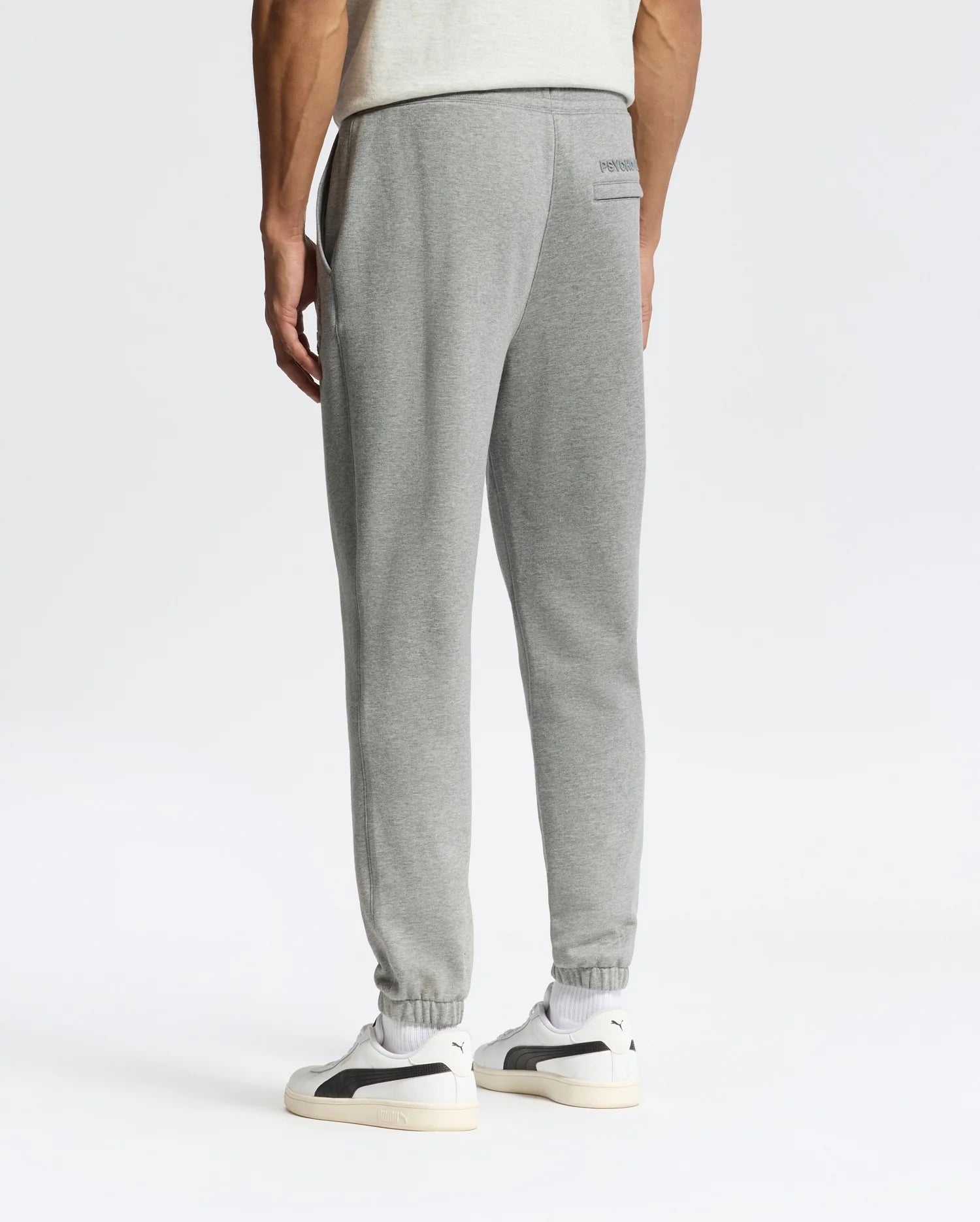 Wearing Psycho Bunny's sleek MENS ELDORADO BRUSH BACK JOGGER - B6P285E200 in gray, a person stands against a plain white background, paired with white sneakers. They face away, highlighting the small logo on the upper back pocket area.