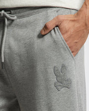 Close-up of someone wearing Psycho Bunny's Men's Eldorado Brush Back Jogger in gray, featuring an embroidered bunny with crossed bones. A drawstring waist provides all-day comfort, and a hand rests casually in the pocket.