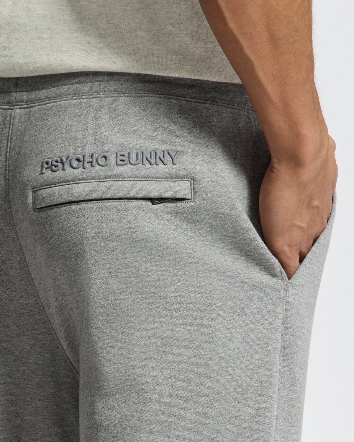 A close-up shows a hand in the pocket of sleek, stylish gray MENS ELDORADO BRUSH BACK JOGGER - B6P285E200 by Psycho Bunny, with "PSYCHO BUNNY" embroidered above the pocket.