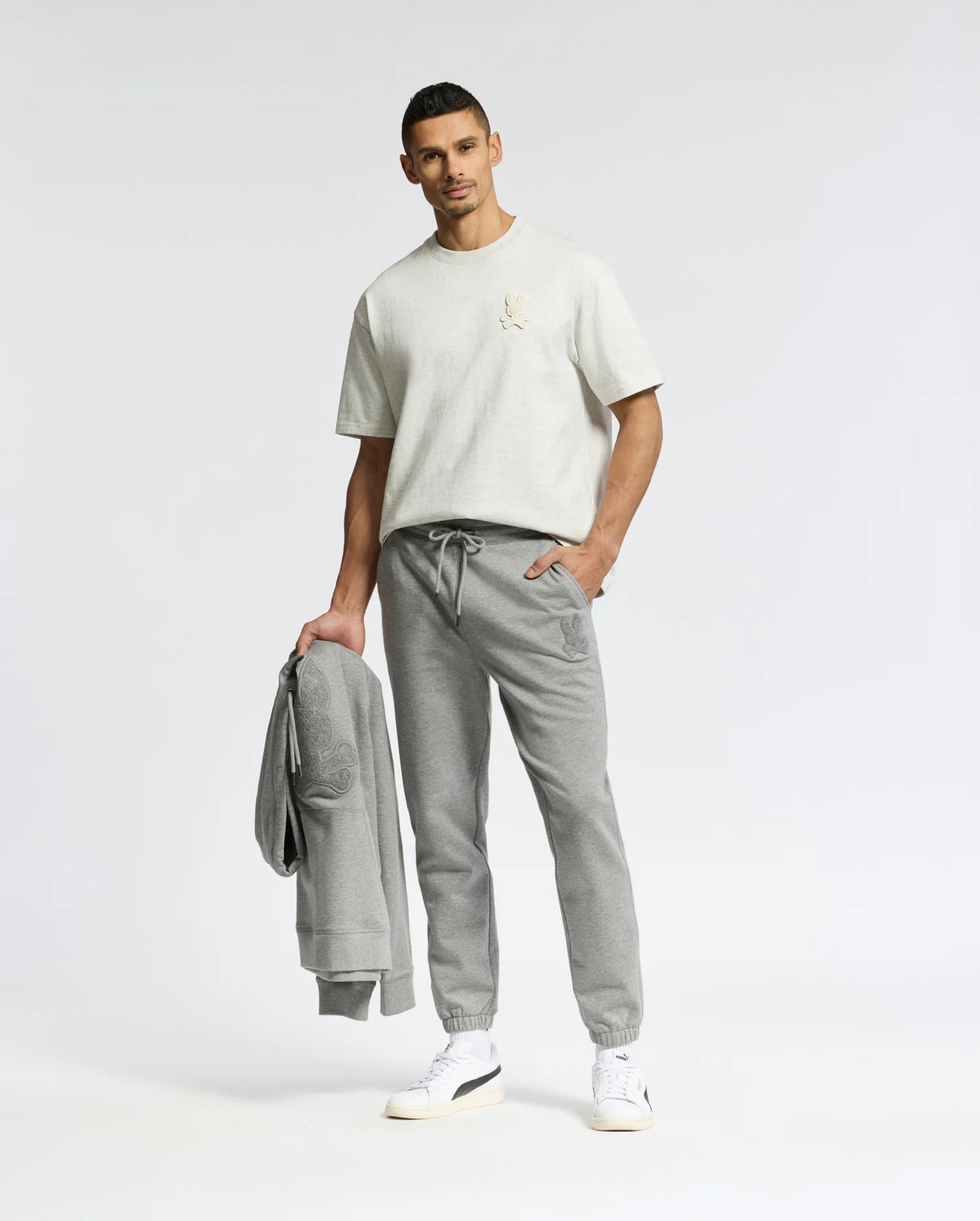 A man stands confidently holding a gray sweatshirt, wearing sleek apparel. His light gray T-shirt pairs perfectly with Psycho Bunny's Eldorado joggers (B6P285E200), while one hand rests in his pocket. White brushback comfort sneakers complete the look against a plain background.