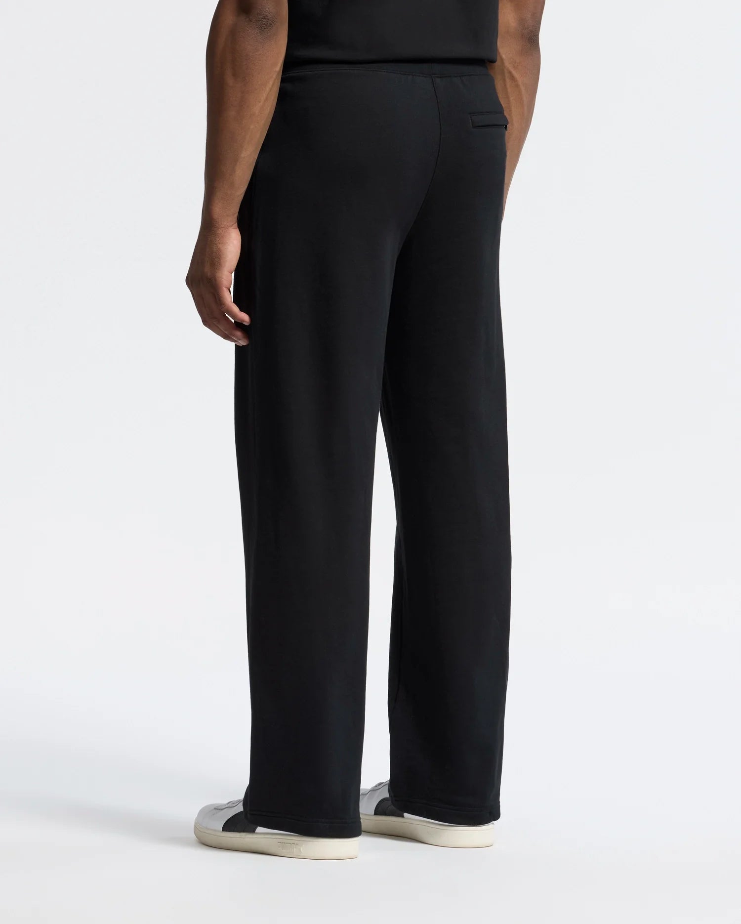 A person with medium skin tone stands back to the camera wearing Psycho Bunny's black MENS LYNX WIDE LEG CLASSIC FRENCH TERRY SWEATPANT and black and white sneakers. Their attire, featuring an embroidered logo, contrasts against a plain white background.