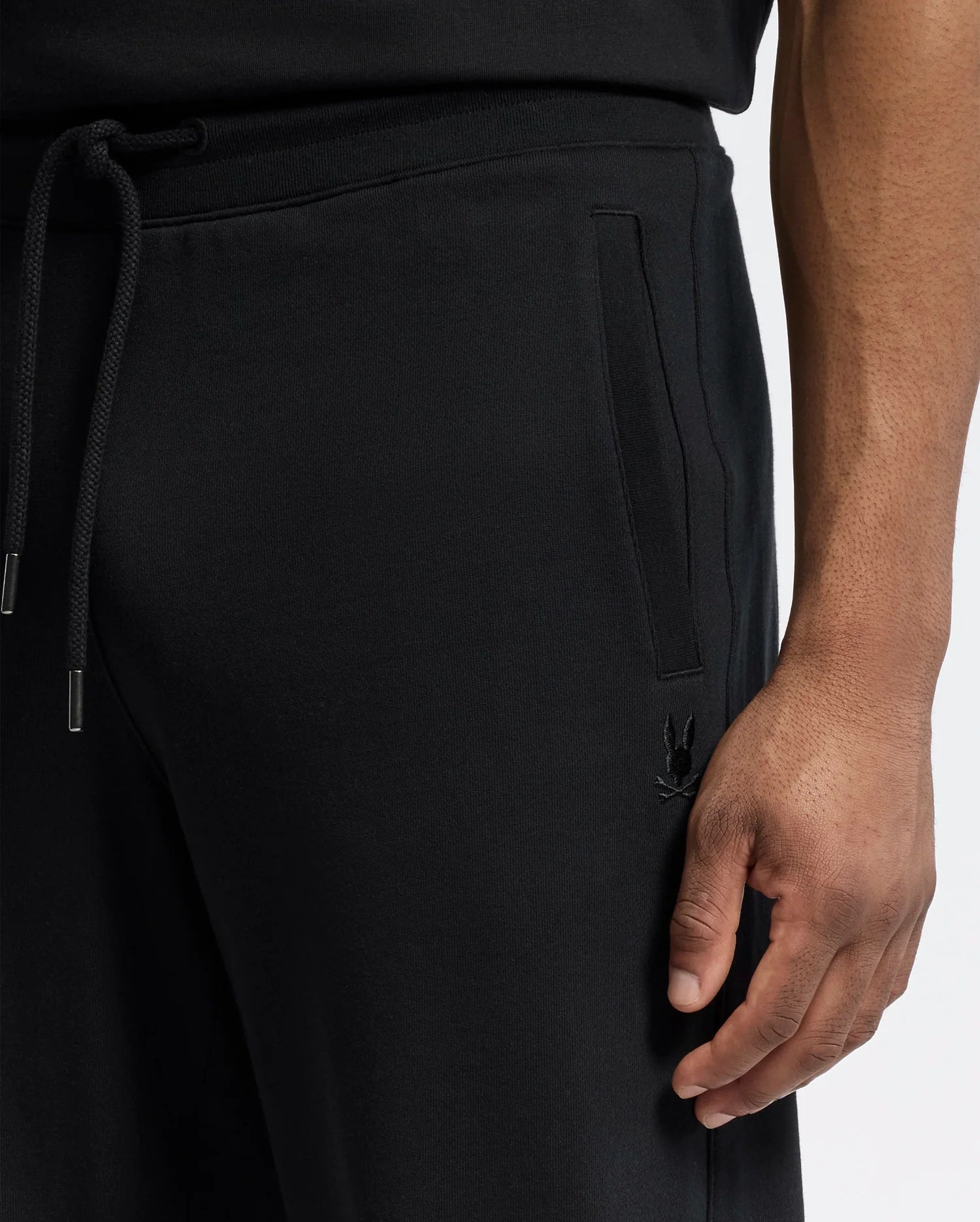 A close-up features a person wearing Psycho Bunny's MENS LYNX WIDE LEG CLASSIC FRENCH TERRY SWEATPANT (B6P379E200) in black with a drawstring and side pockets, an embroidered logo near the pocket, and their right hand relaxed against a plain white backdrop.