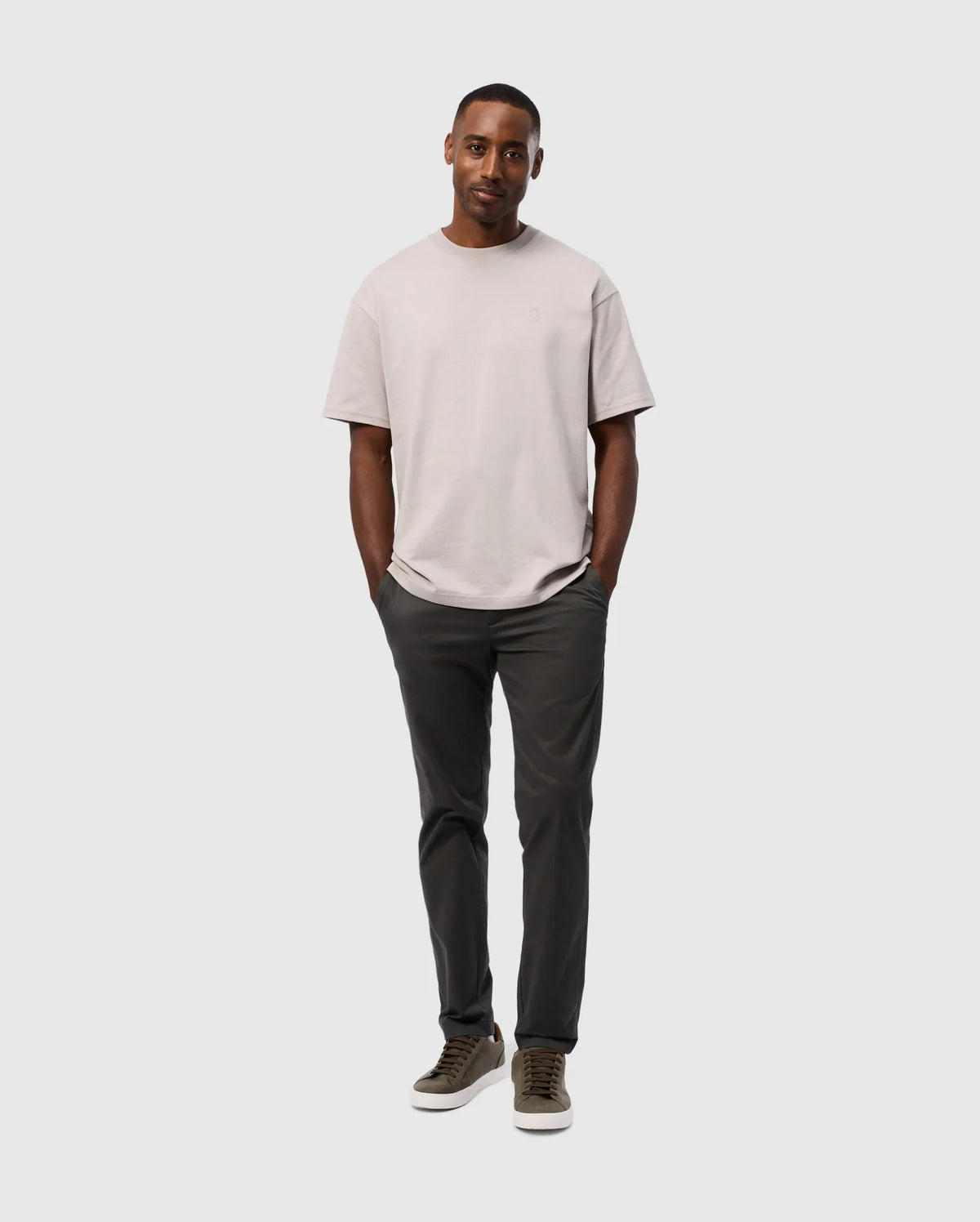 A man stands confidently wearing a beige t-shirt, the MENS GILMAN CHINO PANT - B6P393Z1WB by Psycho Bunny, known for their comfort and stretch, and olive green sneakers. He poses with his hands in his pockets against a light gray background.