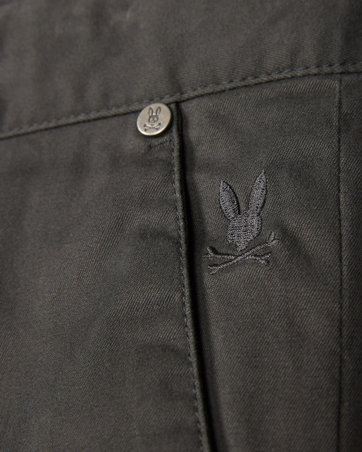Close-up of the MENS GILMAN CHINO PANT by Psycho Bunny, showcasing a dark chino fabric with a pocket. A small, embroidered rabbit skull and crossbones logo adds unique flair, complemented by a metal button embossed with the same rabbit design. The comfortable stretch material ensures both style and ease of movement.