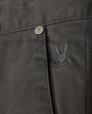 Close-up of the MENS GILMAN CHINO PANT by Psycho Bunny, showcasing a dark chino fabric with a pocket. A small, embroidered rabbit skull and crossbones logo adds unique flair, complemented by a metal button embossed with the same rabbit design. The comfortable stretch material ensures both style and ease of movement.