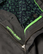 Close-up of the mens Gilman chino pant in a dark color, featuring a zipper and button. The pants have a stretch inner lining adorned with green bunny patterns on a navy background. To enhance comfort, the waistband is embroidered with the brand name "Psycho Bunny" in green.