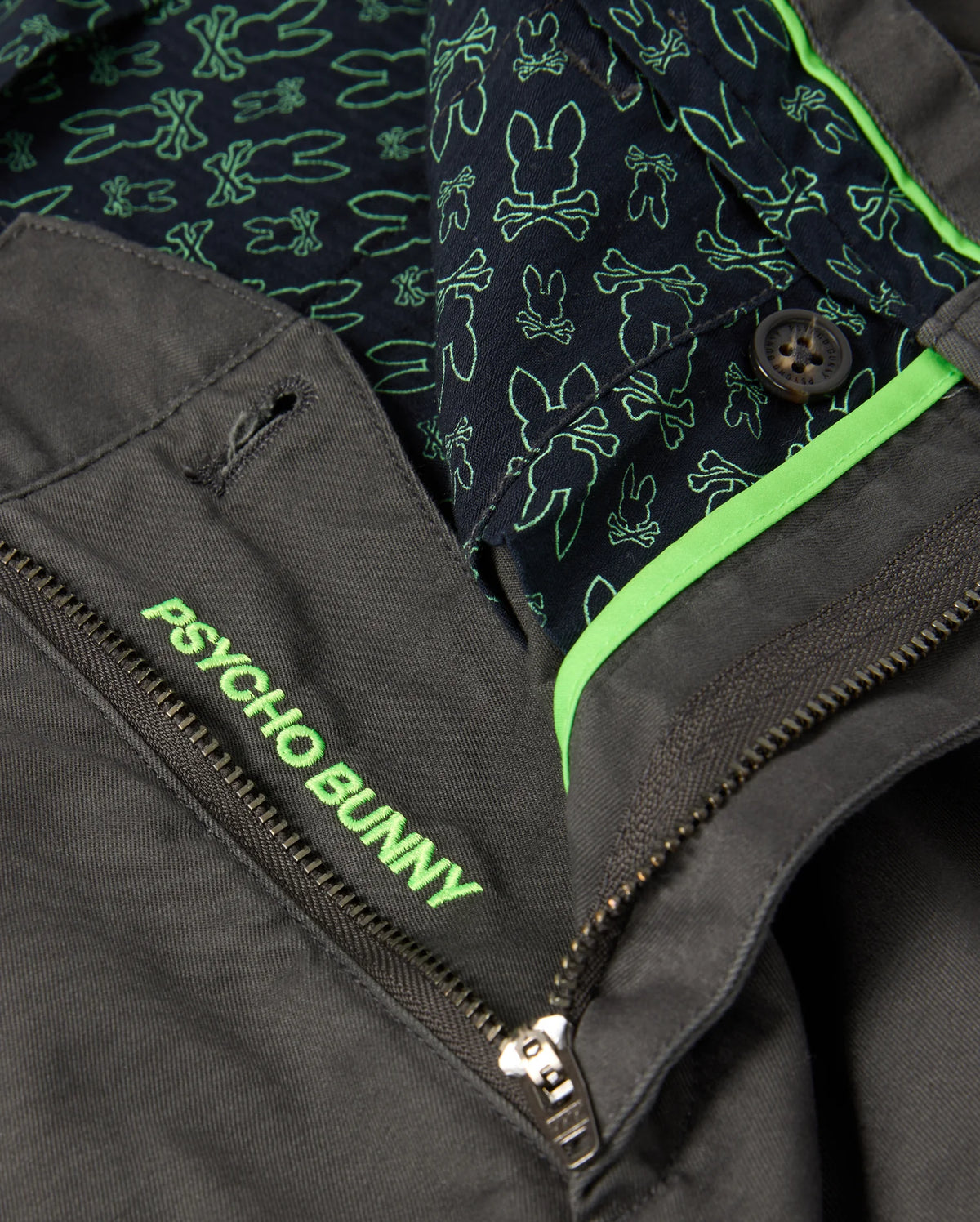 Close-up of the mens Gilman chino pant in a dark color, featuring a zipper and button. The pants have a stretch inner lining adorned with green bunny patterns on a navy background. To enhance comfort, the waistband is embroidered with the brand name "Psycho Bunny" in green.